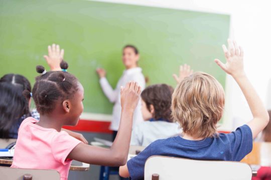 Young students sitting at their desks in a classroom with their hands raised