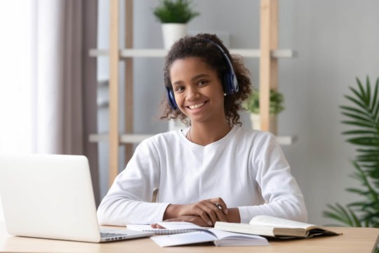 Young girl smiling and sitting at a desk with a laptop and open book.