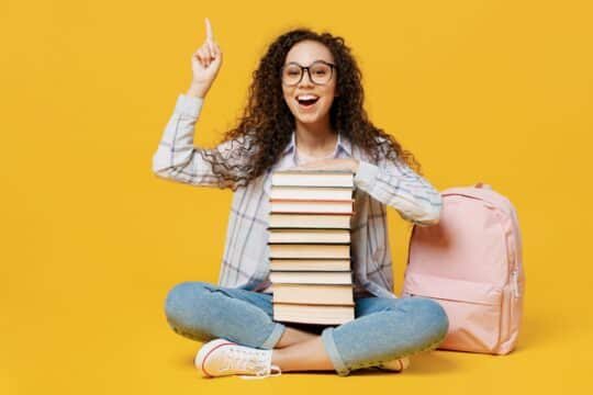 A girl sits on the ground with books and a pencil, smiling.