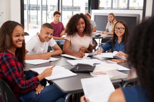 A group of high school students sitting around a table listening to their teacher