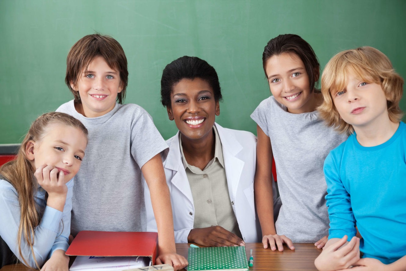 Female teacher and a group of students smiling in front of chalkboard