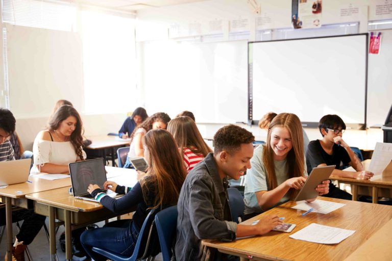 Classroom full of students working at tables together with papers, tablets, and laptops.