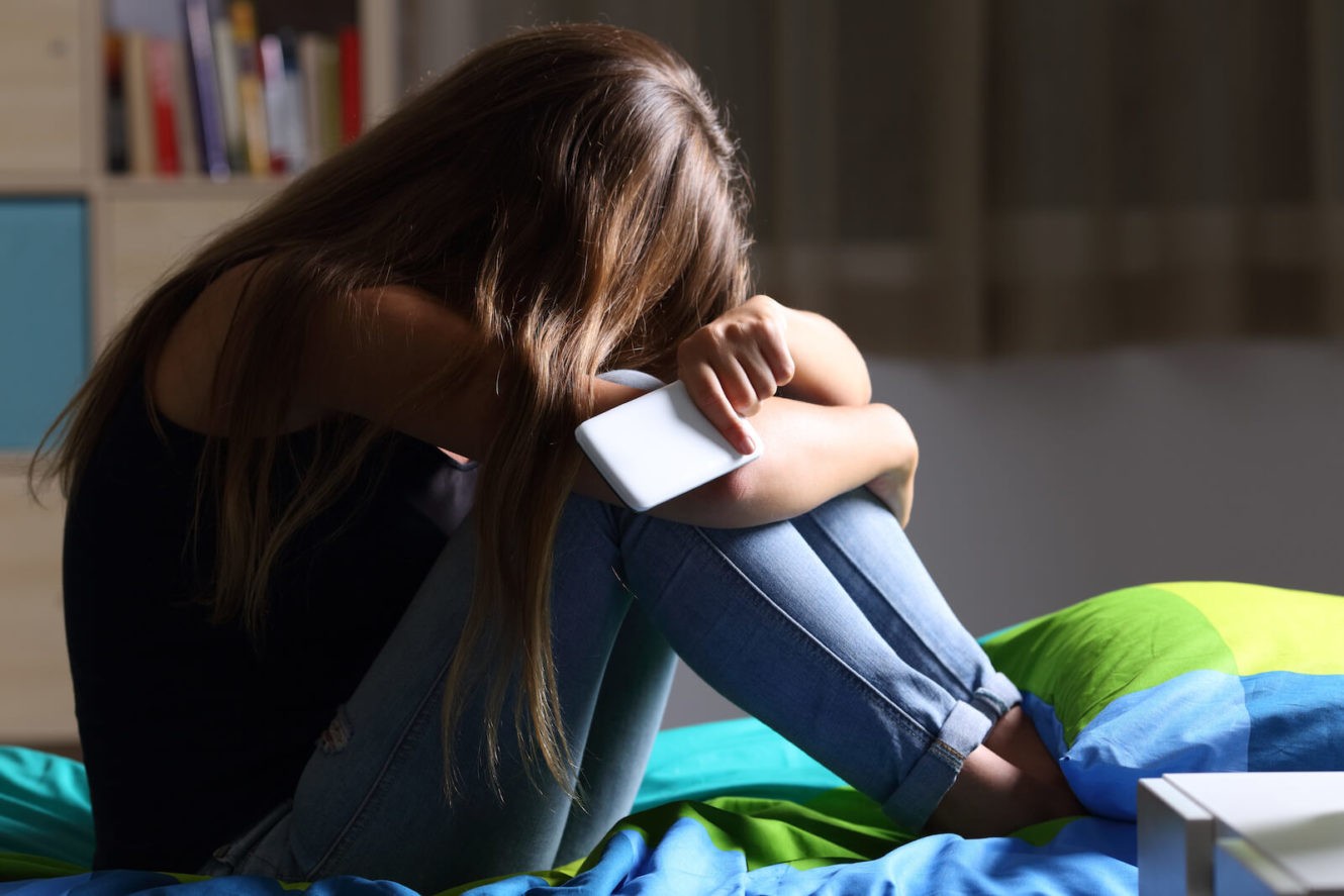 Sad girl sitting on her bed with her head down while holding her cell phone