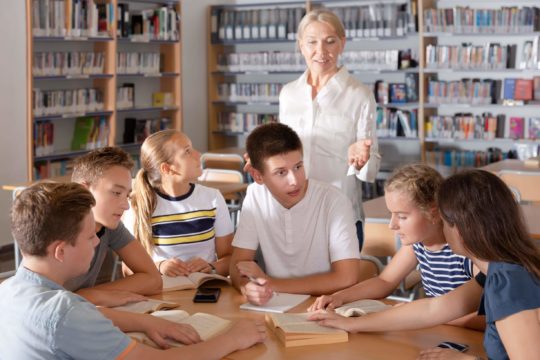 A group of high school students sit in a group with books, discussing with the help of a teacher.