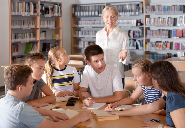 A group of high school students sit in a group with books, discussing with the help of a teacher.