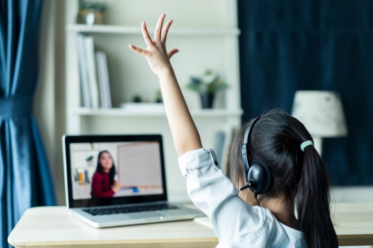 Young girl raising her hand while watching her teacher in a Zoom class.