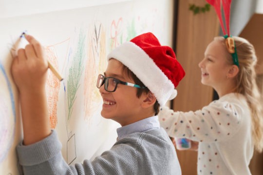 Two students wearing Santa hats drawing on paper hanging on a wall.