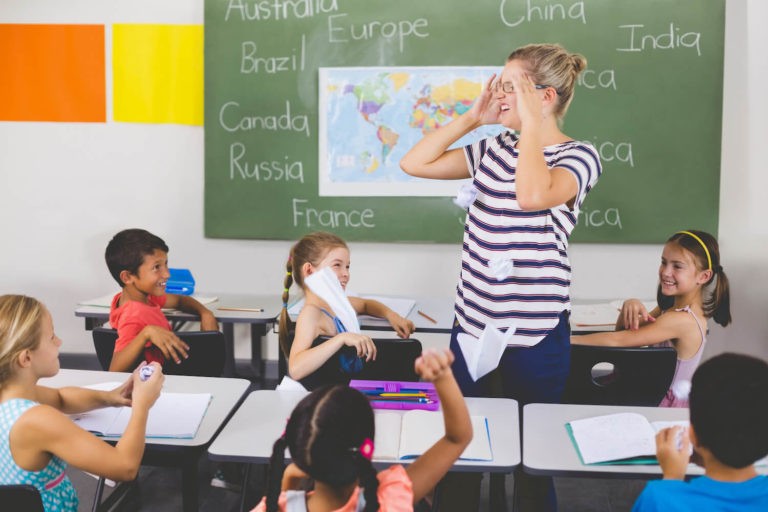 Frustrated teacher holding her head with a chaotic classroom of students.