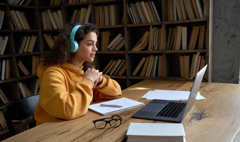 Girl with headphones on watching a Zoom class while taking notes.