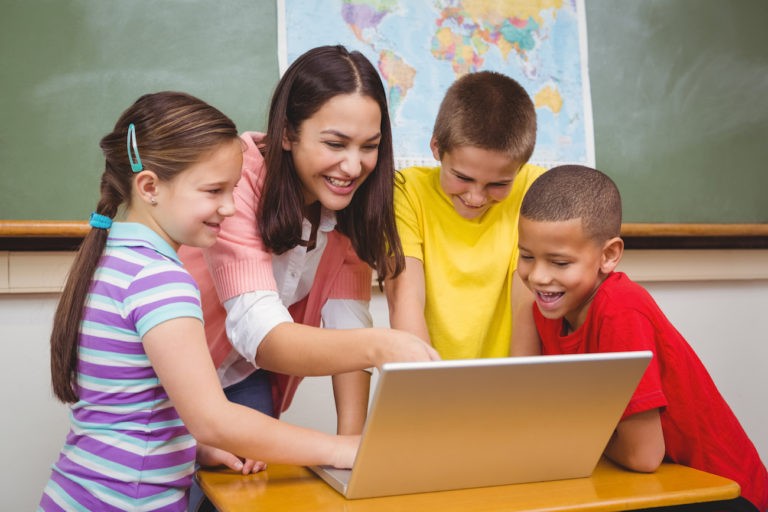 A teacher and group of young students standing in a classroom using a laptop.