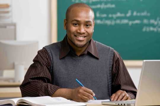 Young teacher sitting in a classroom at a desk with a laptop and grading papers