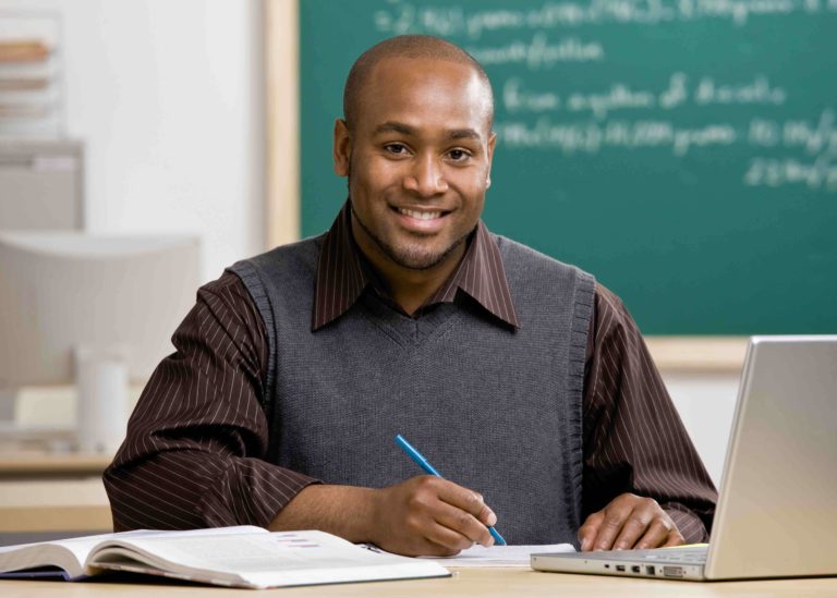 Young teacher sitting in a classroom at a desk with a laptop and grading papers