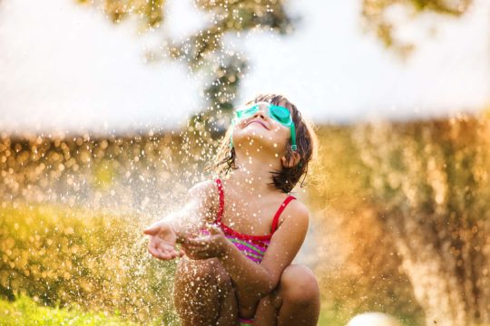 Little girl in a bathing suit and goggles playing outside in a sprinkler.