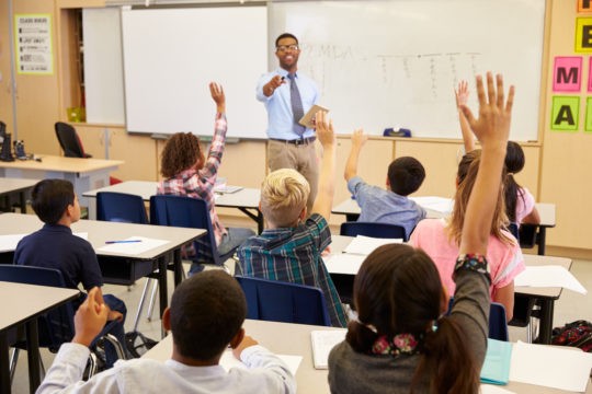 Teacher standing at the front of a class pointing to a student with their hand up.