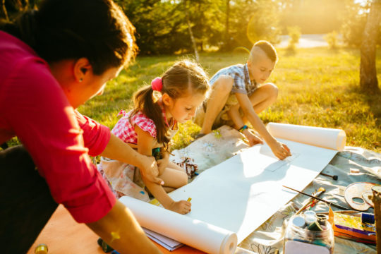 An adult and two kids outside drawing and painting on paper.