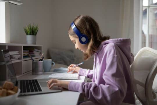 A girl sits at her desk with headphones on, listening to a podcast.