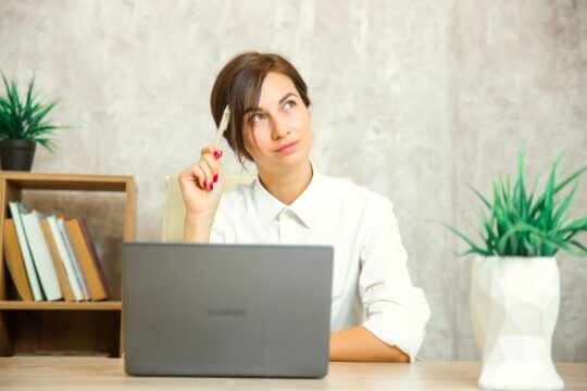 A woman sits at her laptop, appearing thoughtful.