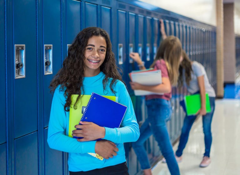 Teenage girl smiling and holding notebooks in a school hallway.