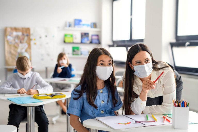 Masked teacher pointing something out while helping a masked student.