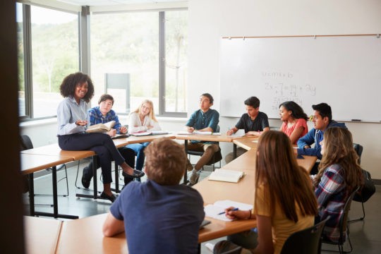 Math teacher talking to students sitting at desks in a half circle.