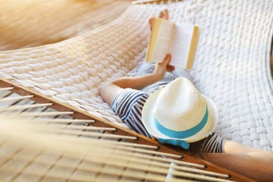 A man lays on a hammock in the sun, reading a book.