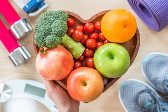 Heart-shaped bowl of colorful fruits and vegetables surrounded by fitness items.