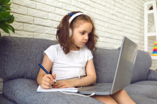 Young student sitting on couch with laptop and notebook while wearing headphones