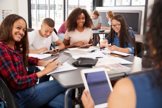 Teacher sits with high school students as they all use tablets to do school work.