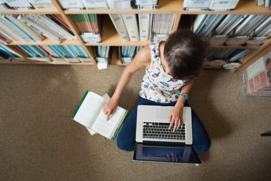 Girl sitting on the floor of a library reading a book and using a laptop.