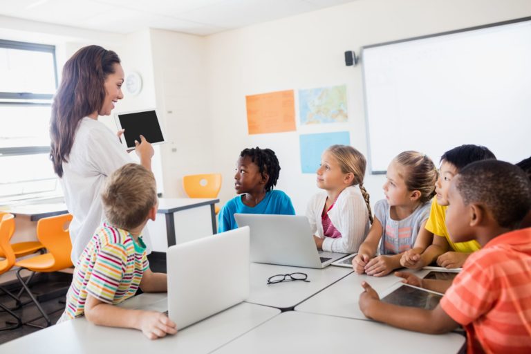 Group of students seated around a table while a teachers shows something on a tablet.