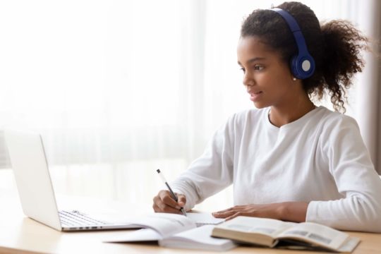 Young girl writing notes while looking at a laptop with open books around her.