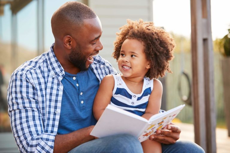 A father and young daughter sitting together outside reading a book.