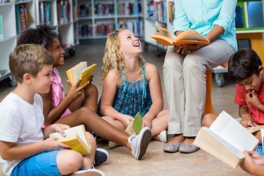 A group of young smiling students reading along to a book with their teacher