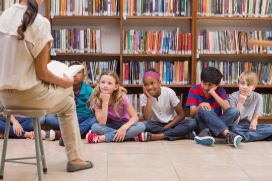 A school librarian reads a book to a group of children in the library.