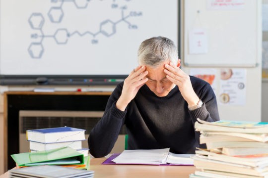 A stressed science teacher tries to grade homework in a classroom.