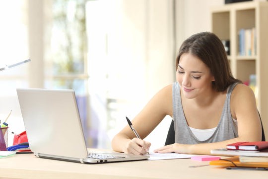 Female high school student writing on paper in front of laptop