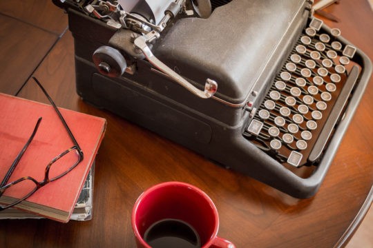 Black typewriter sitting on a table with books, glasses, and coffee